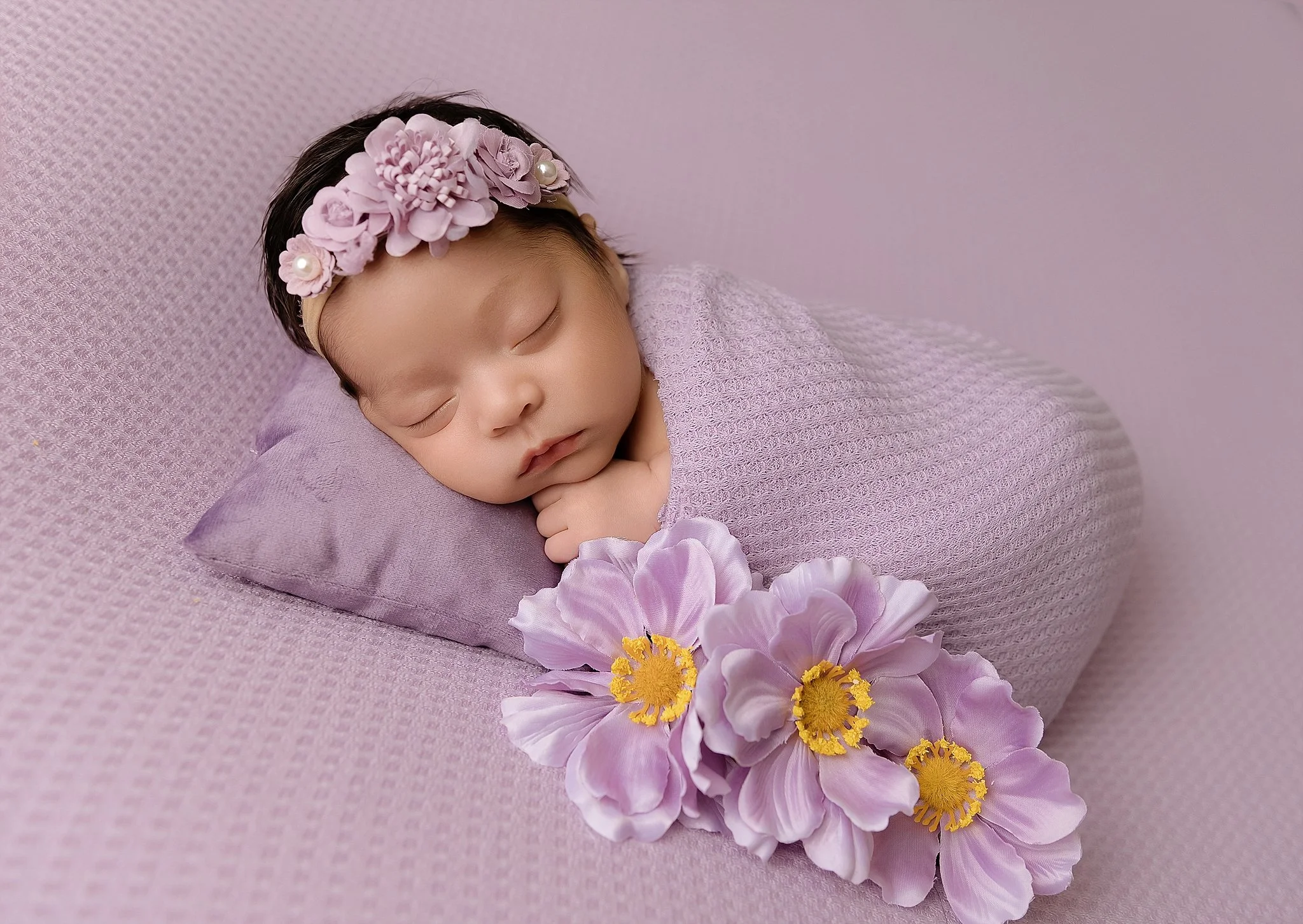 Sleeping newborn baby girl wrapped in soft lavender fabric with a floral headband, photographed in a home studio in Elk Grove, California.