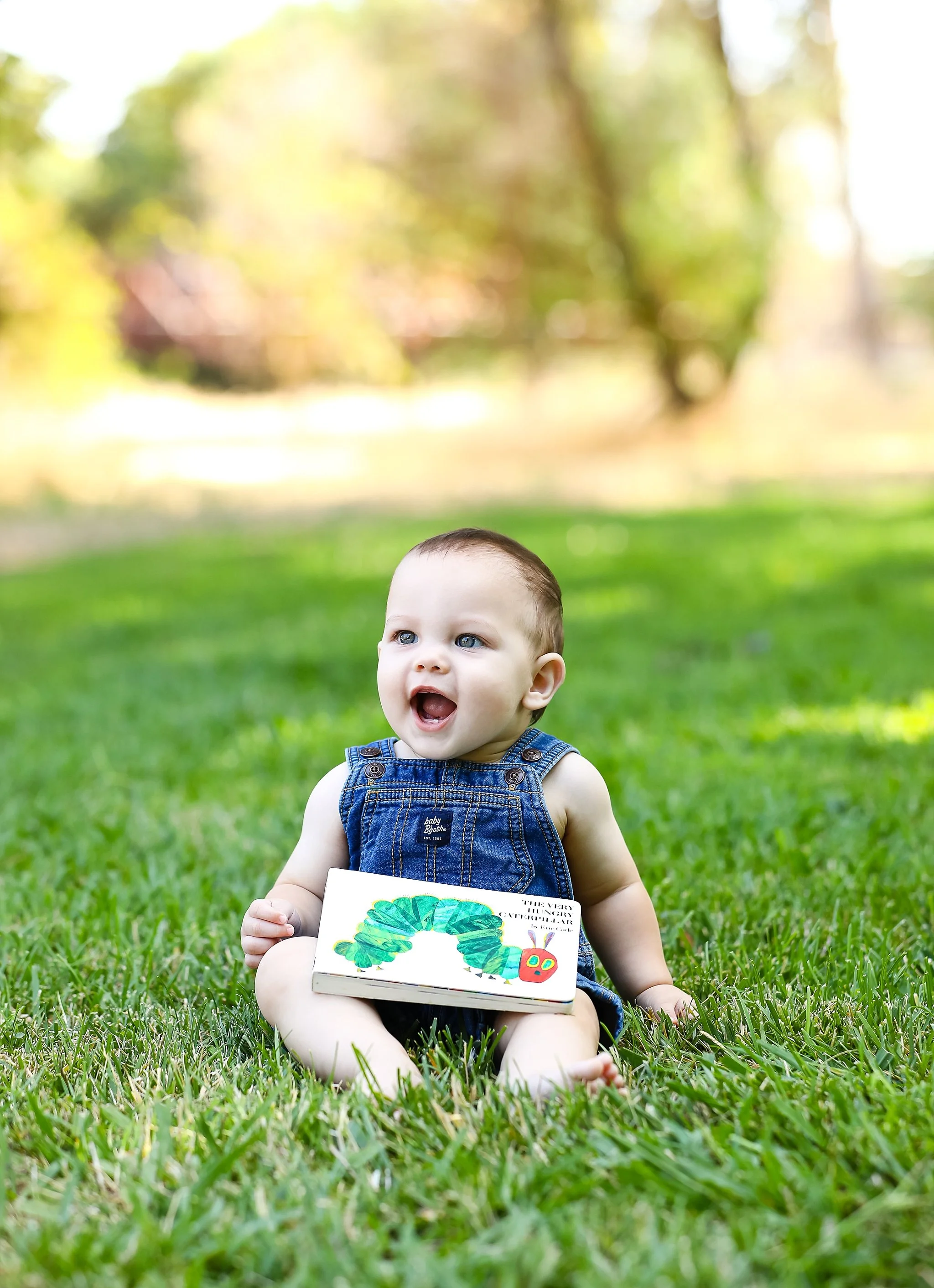 Nine-month-old baby boy sitting in the grass holding a favorite children's book during an outdoor milestone photography session in Elk Grove, California.