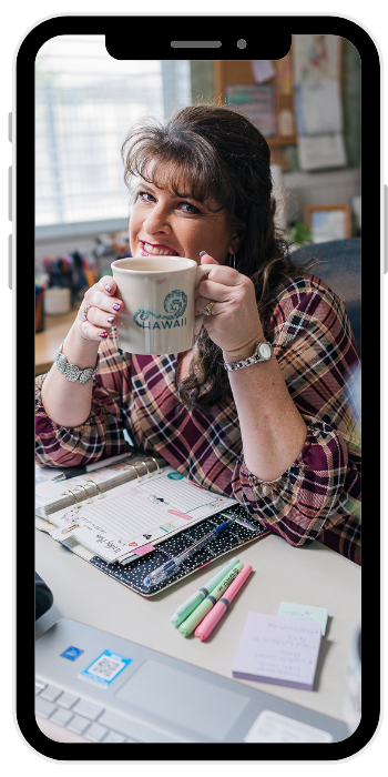 A woman with brown hair and bangs smiling and holding a white mug with the word 'Hawaii' and a seahorse graphic, sitting at a desk with notebooks, pens, and sticky notes in an office or home workspace.