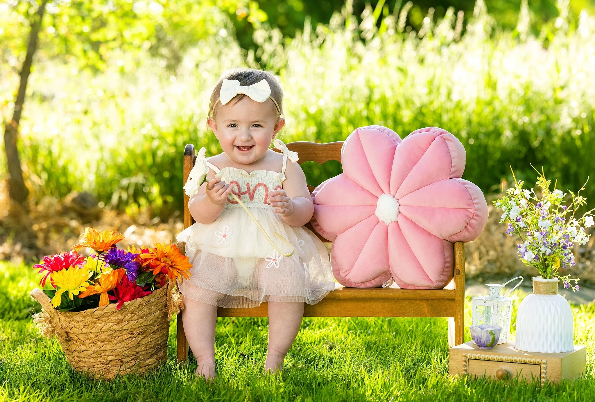 Smiling baby girl sitting on a wooden bench with colorful spring flowers during a milestone session in Elk Grove, California.