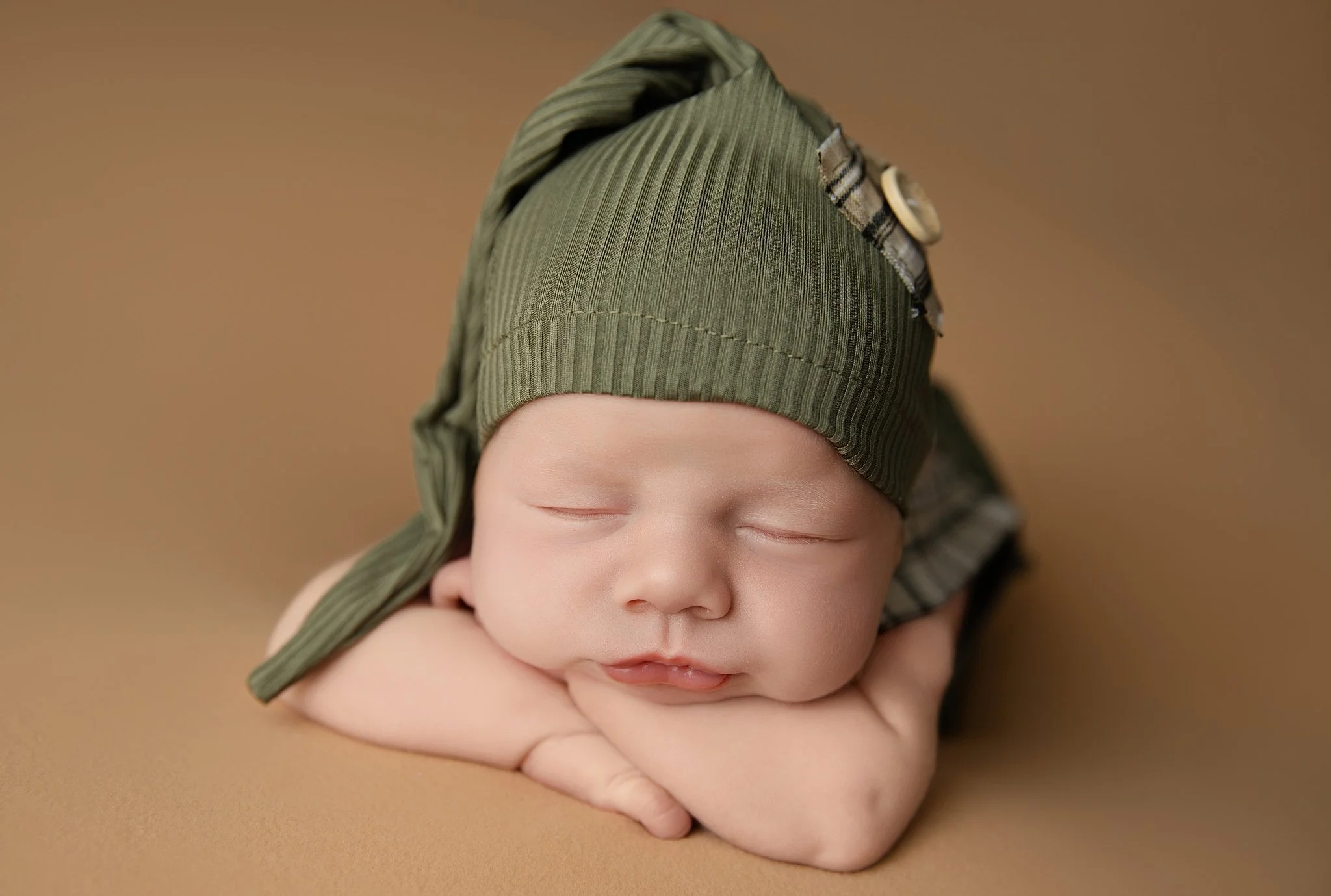 Close-up portrait of a sleeping newborn baby boy wearing a green bonnet against a warm neutral background in an Elk Grove home studio.