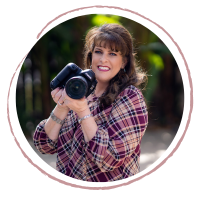A woman holding a professional camera, smiling outdoors in a natural setting.