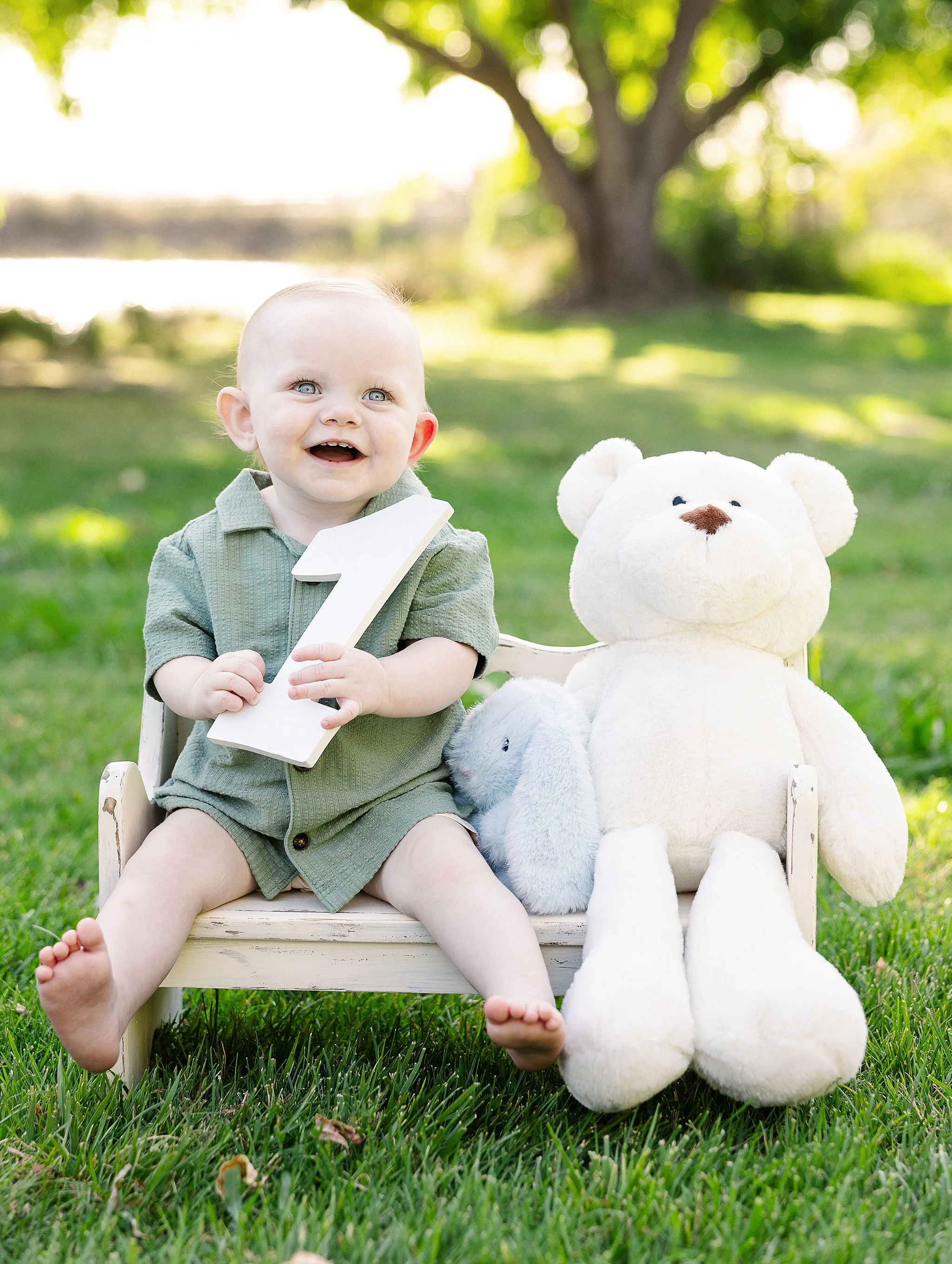 Smiling one-year-old boy holding a wooden number one while sitting beside a teddy bear during an outdoor milestone portrait session in Elk Grove, California.