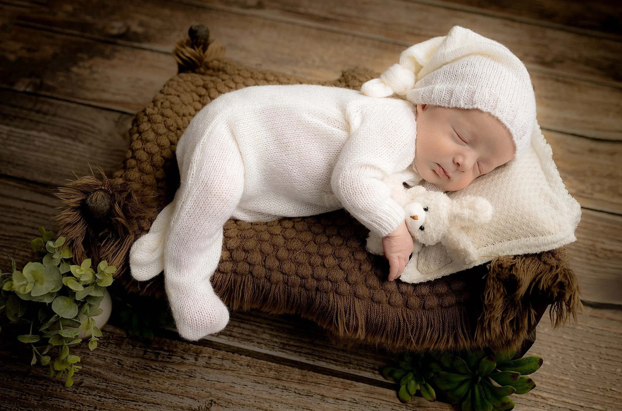 Sleeping newborn baby in a cream knit outfit and bonnet posed on a rustic wooden prop in an Elk Grove newborn photography studio.