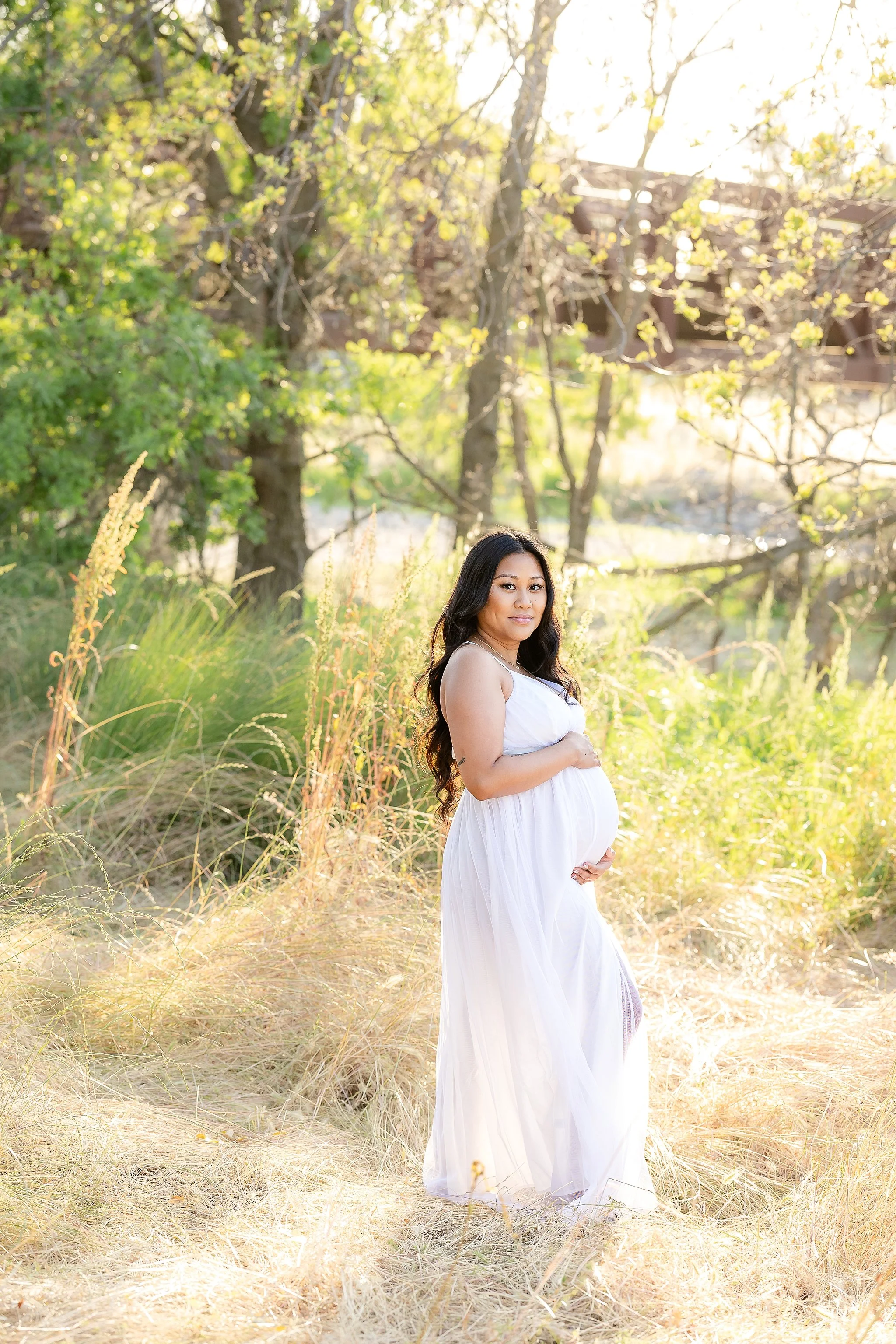 Outdoor maternity portrait of an expecting mother in a white dress holding her baby bump during golden hour in Elk Grove, California