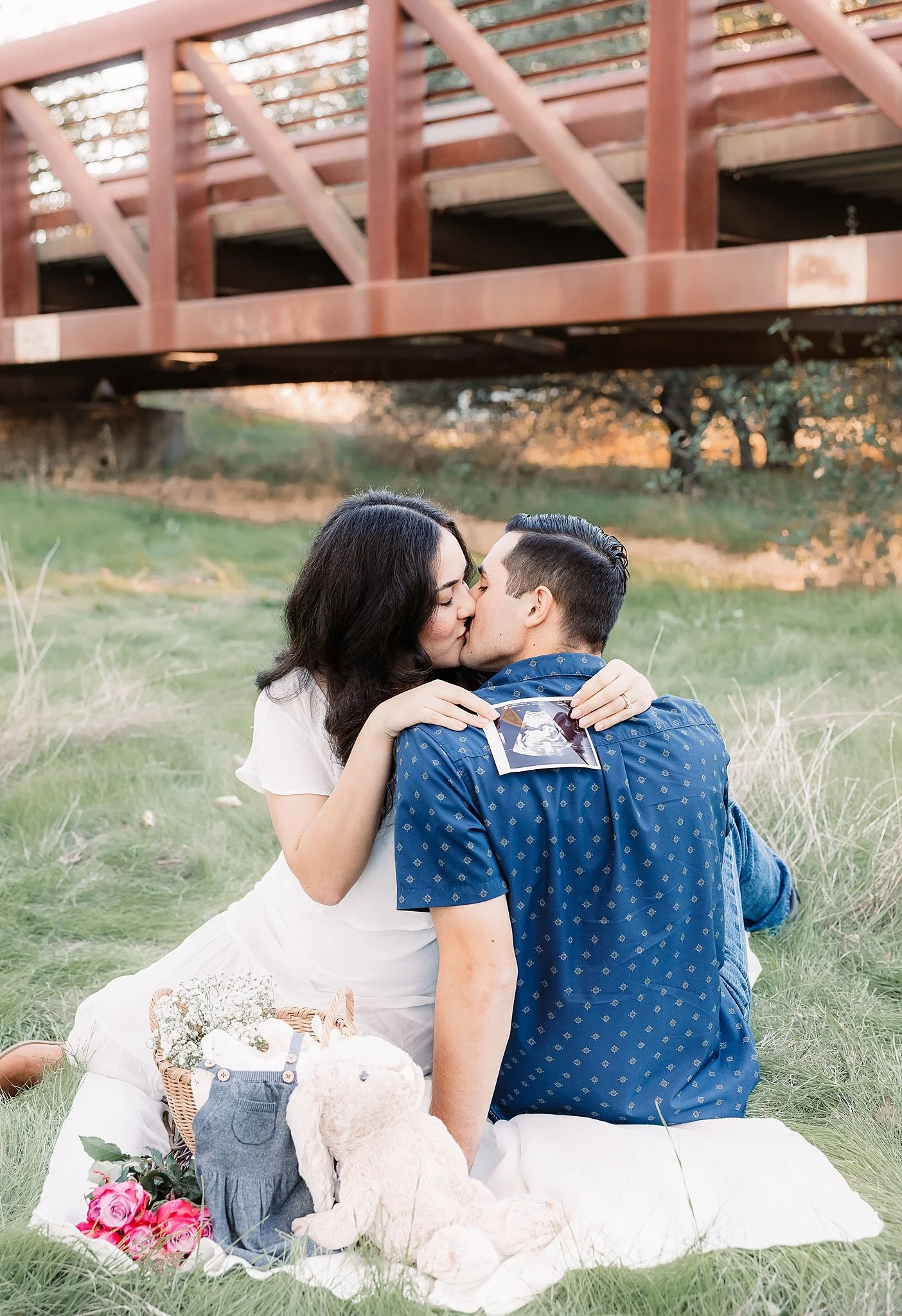 Outdoor maternity session in Elk Grove, California featuring an expecting couple sharing a kiss with an ultrasound photo and baby props displayed beside them