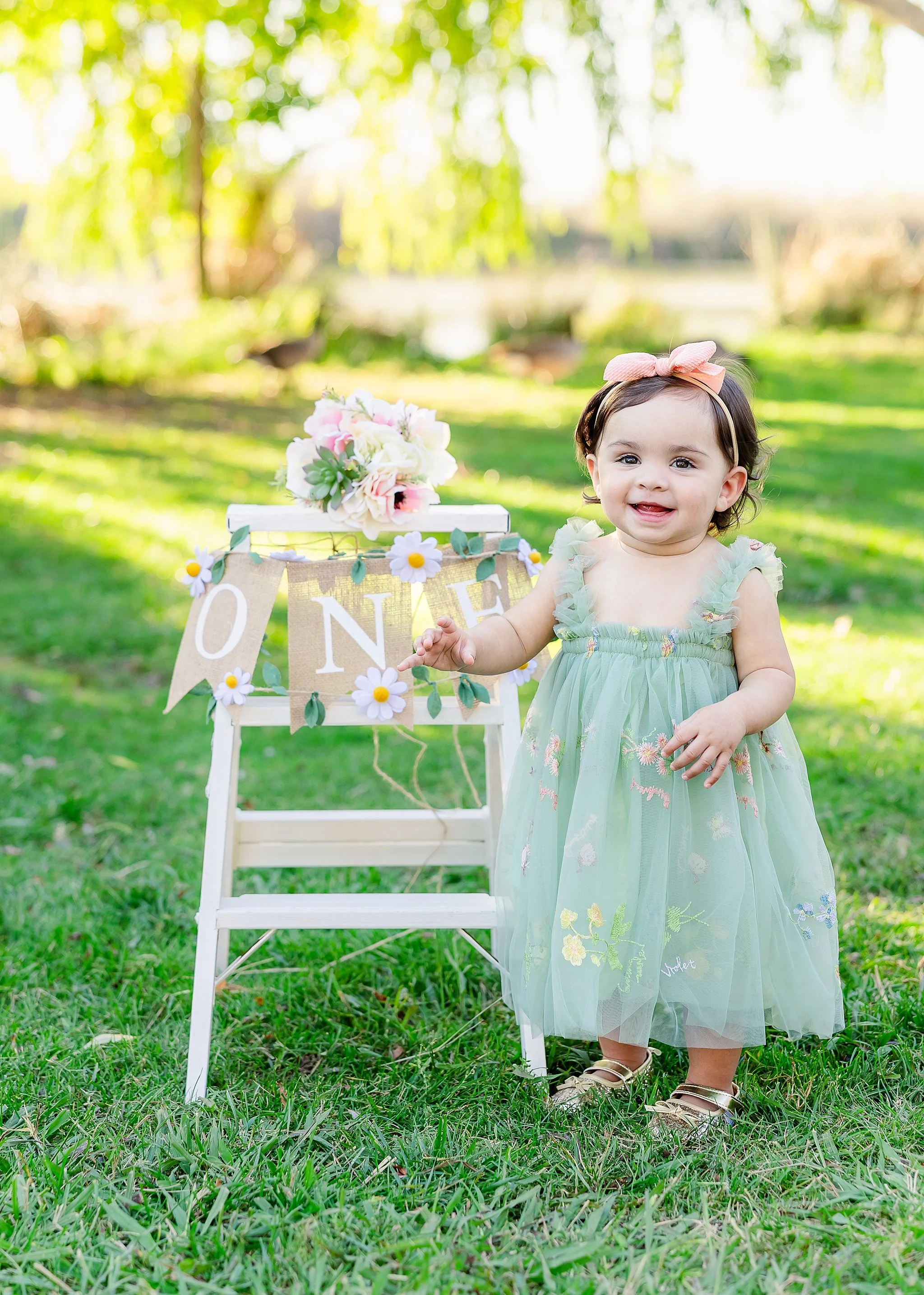 One-year-old girl standing beside a floral-decorated stool during an outdoor first birthday milestone portrait session in Elk Grove, California.