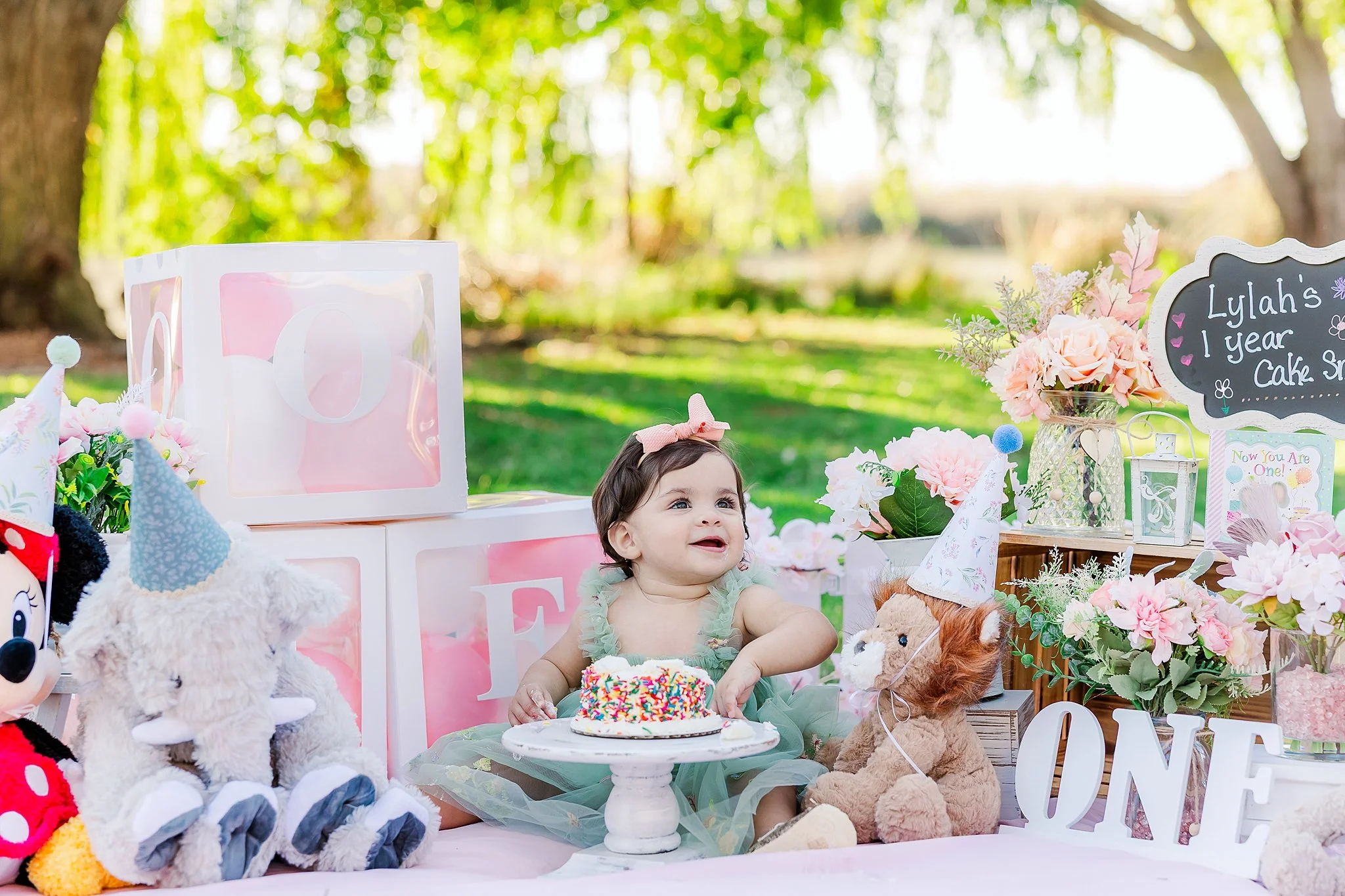One-year-old girl enjoying her cake smash surrounded by flowers and stuffed animals during an outdoor first birthday photography session in Elk Grove, California.