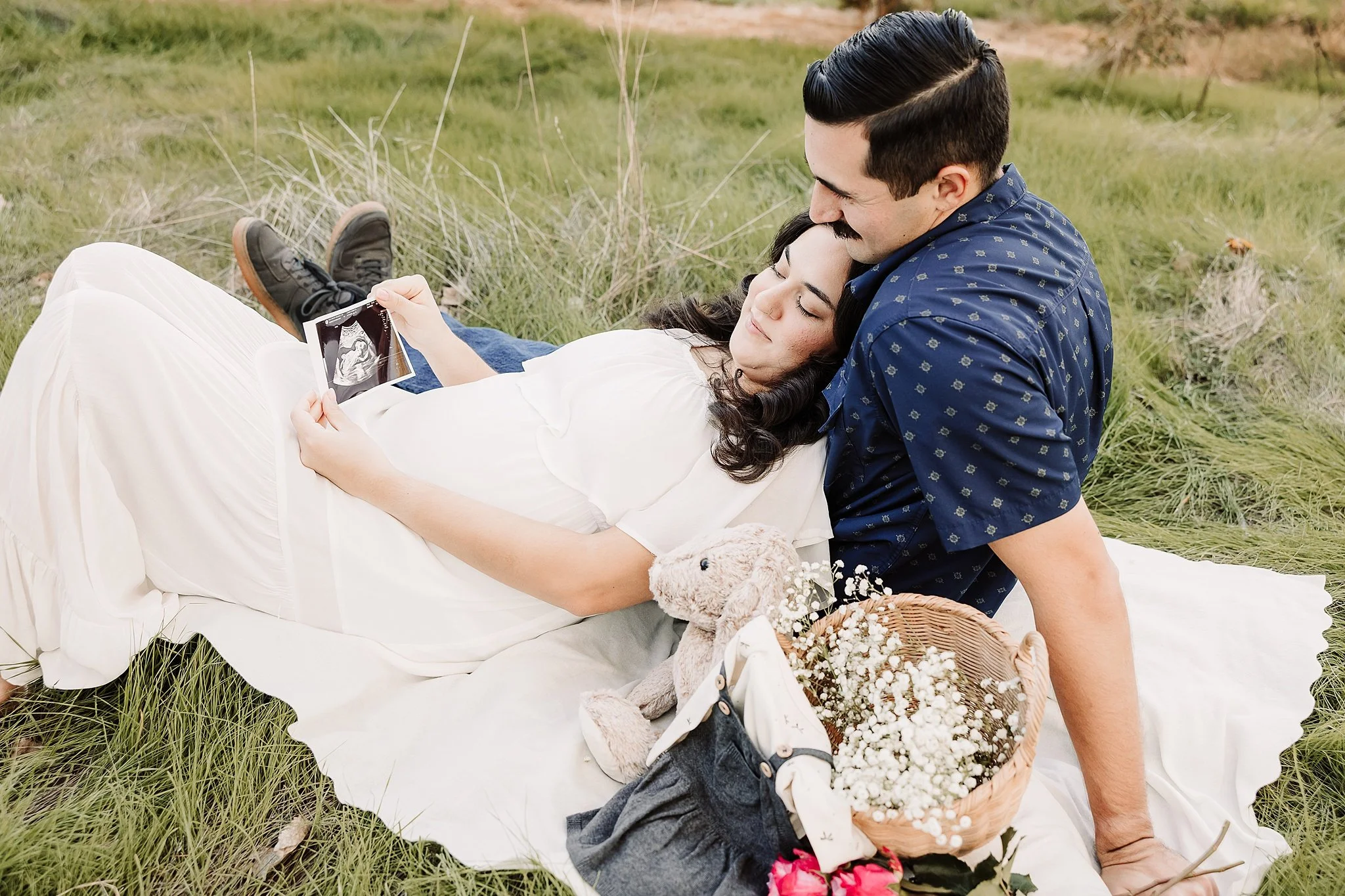 Expecting couple relaxing on a blanket during an outdoor maternity session, holding an ultrasound photo with baby outfit and flowers nearby in Elk Grove, California