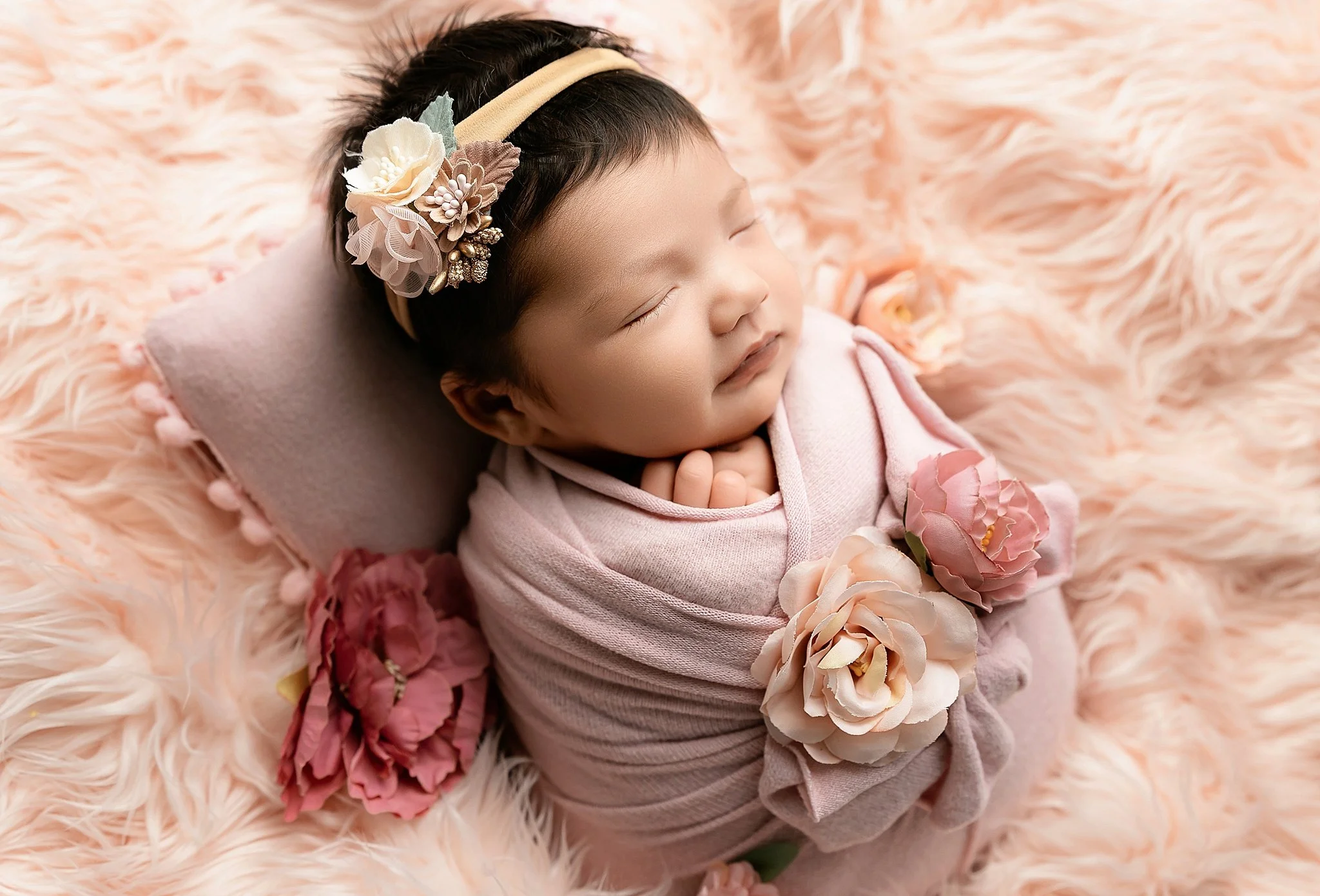 Newborn baby girl wrapped in blush fabric with floral accents posed on a soft peach backdrop in Elk Grove, CA.