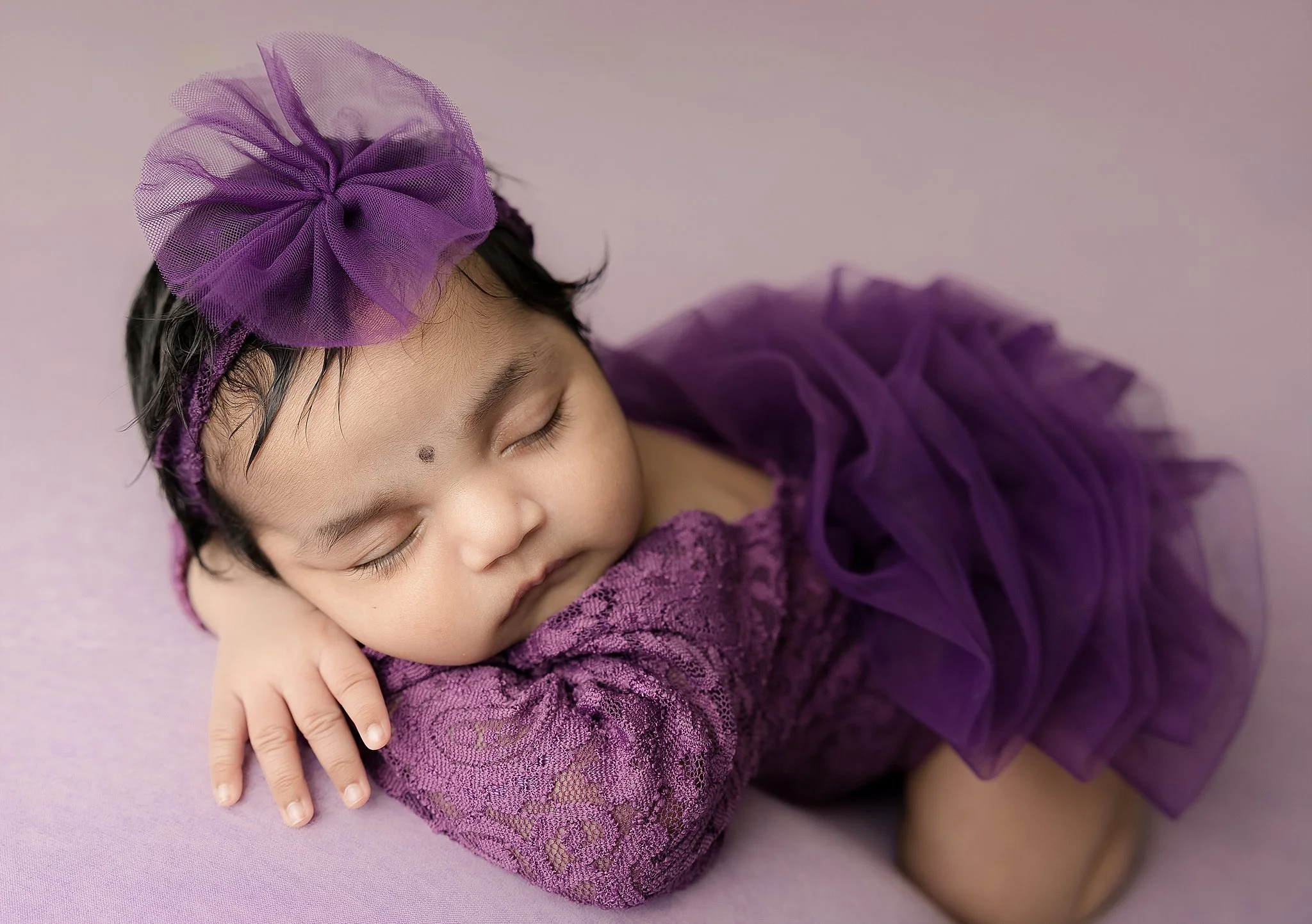 Sleeping newborn baby girl wearing a deep purple lace outfit and bow headband, posed on a soft backdrop in an Elk Grove newborn studio.