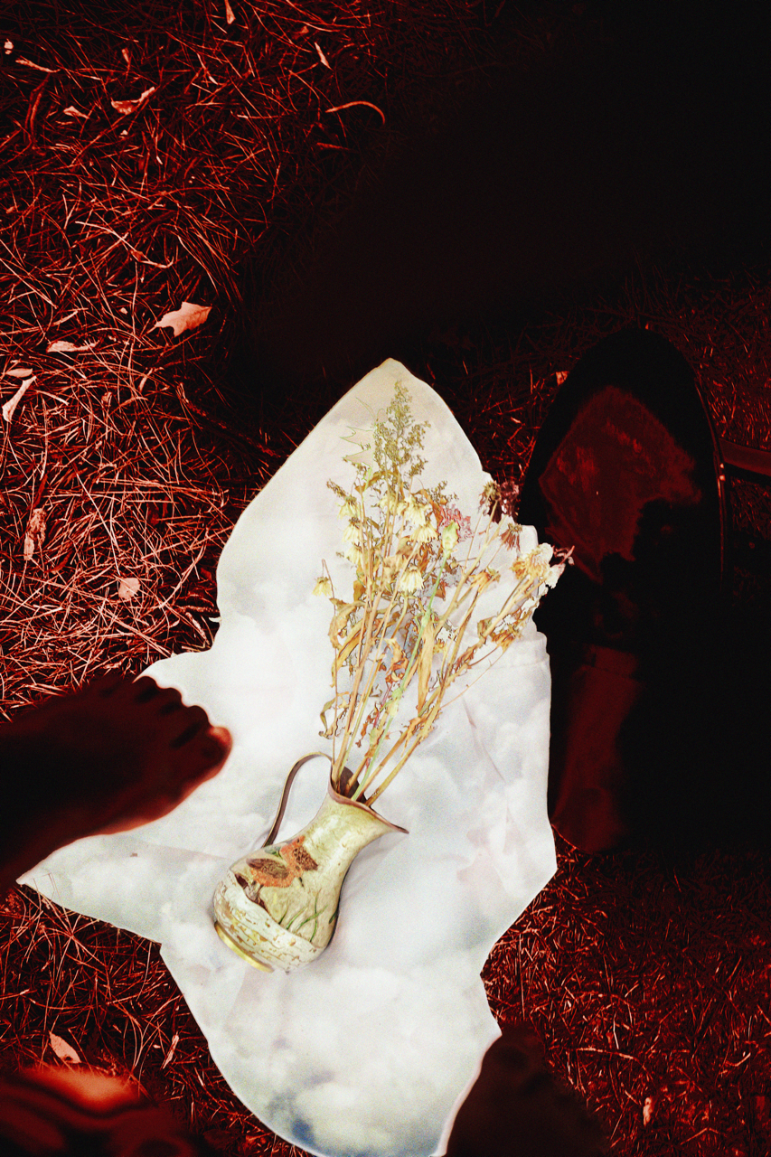 A person holding a piece of paper with a picture of a vase with dried flowers on it, surrounded by grass and foliage with a black object lying nearby.