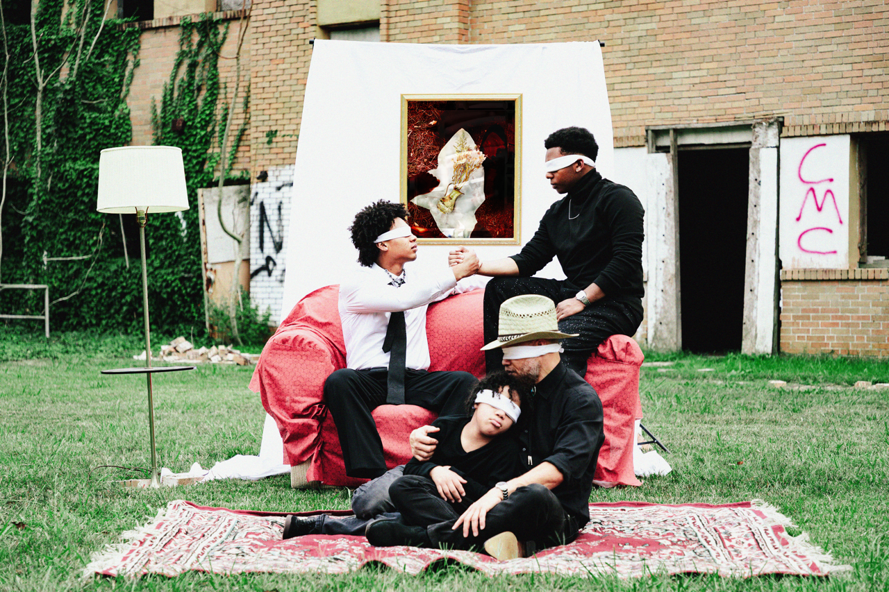 Four men with white blindfolds in an outdoor setting with a makeshift art installation that includes a sofa, a painting, and a rug, in front of a brick building with graffiti.