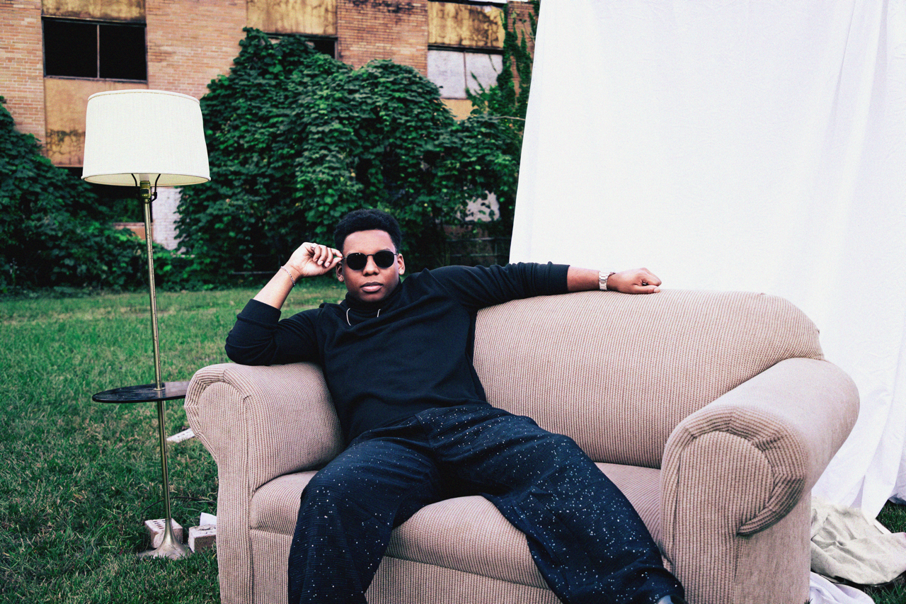A young man sitting on a pink sofa outdoors, wearing sunglasses, a black long-sleeve shirt, and black pants with white paint splatters, with a white curtain to his right and a lamp standing next to the sofa.
