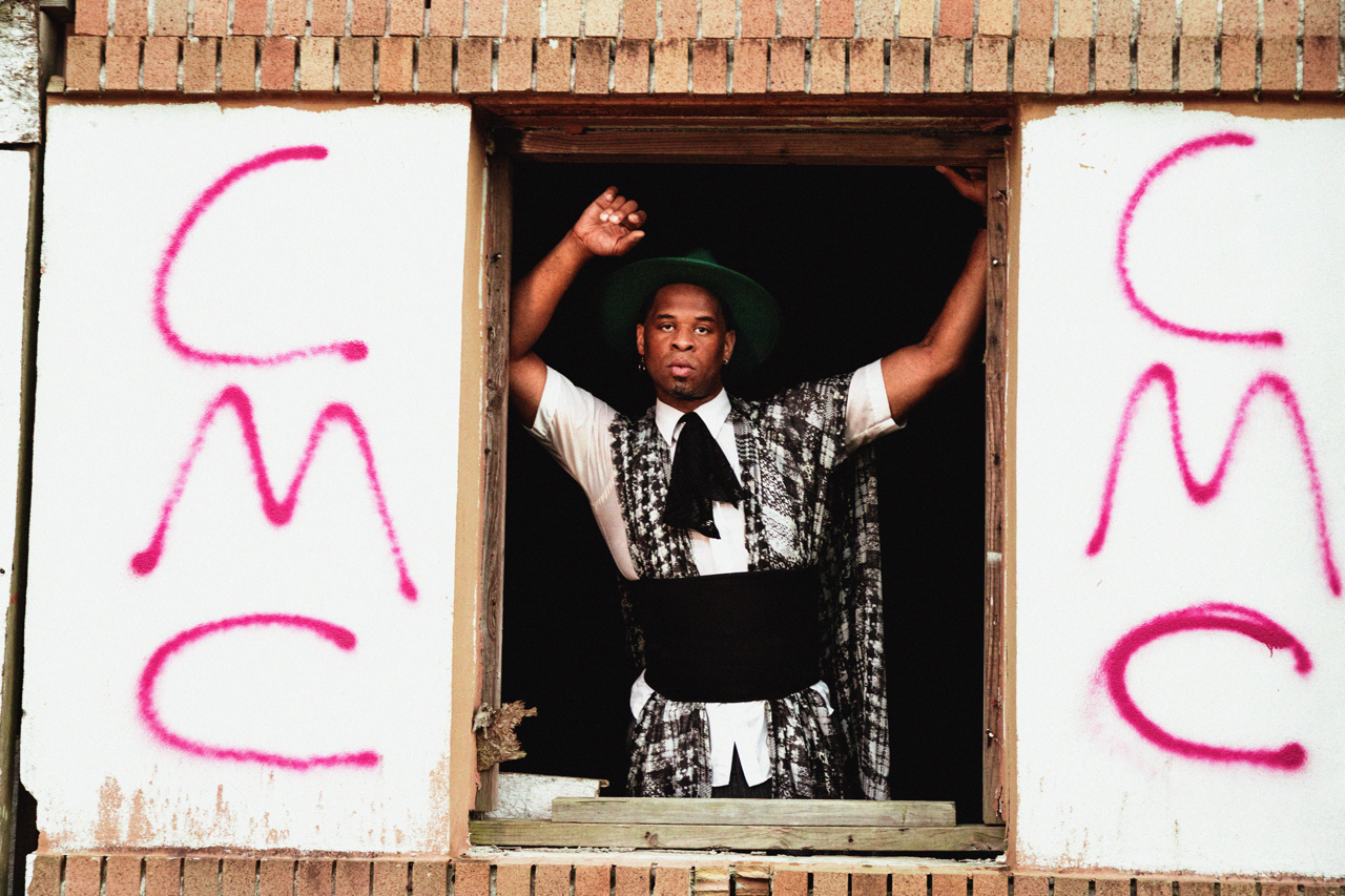 A man standing in an unfinished window, wearing a patterned outfit with a black wrap-around waist, a white shirt, and a black necktie, also wearing a green wide-brimmed hat, with graffiti on the white wall on either side spelling out "CAME" in pink s