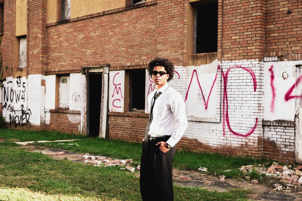 A man in a white shirt, black pants, and sunglasses standing in front of an abandoned building with graffiti on the walls.
