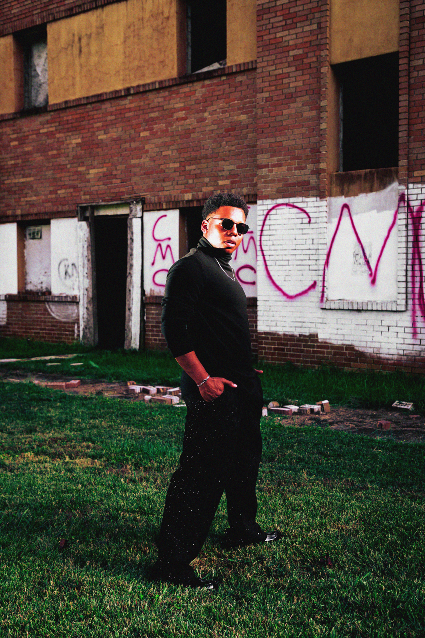 Young man standing on grass in front of a graffiti-covered building, wearing sunglasses, a black turtleneck, and black pants.