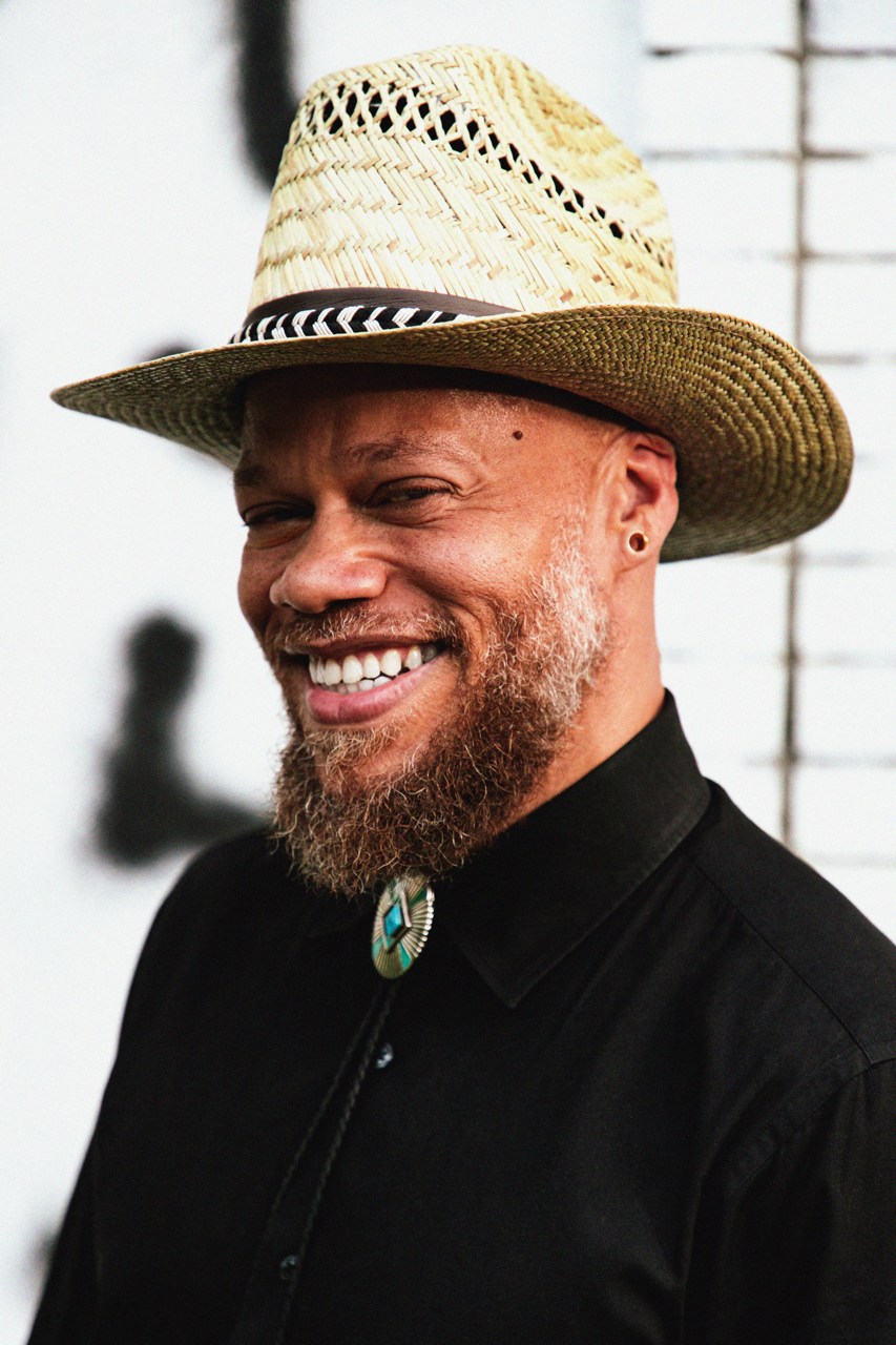 A smiling man wearing a light-colored straw hat with black and white striped band, a black shirt, and a bolo tie, standing in front of a white wall with some black abstract art.