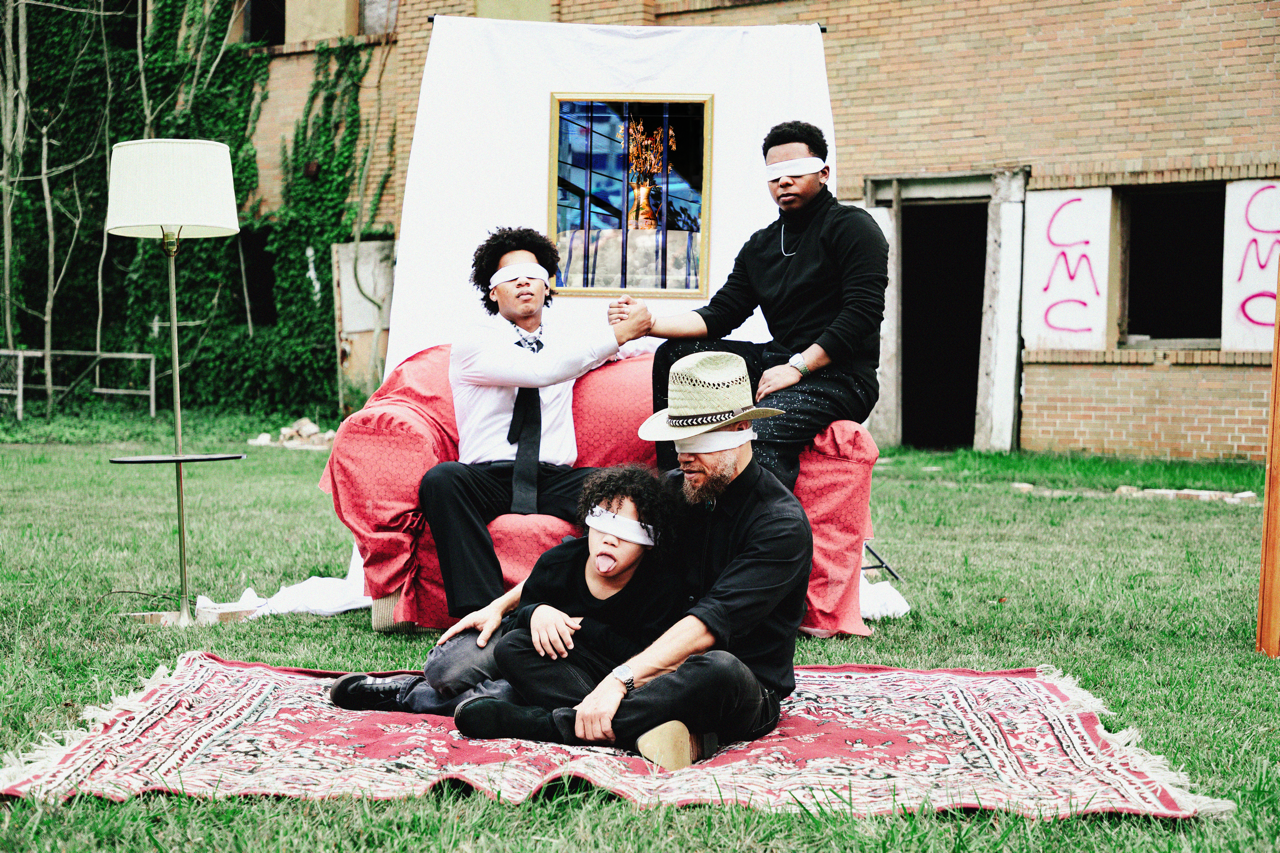 Five people outdoors on a grassy lawn, all wearing eye masks. One person is sitting on a red chair, one on the ground, and three sitting on a large cushioned surface. They appear to be engaged in a playful or artistic activity, with a background that