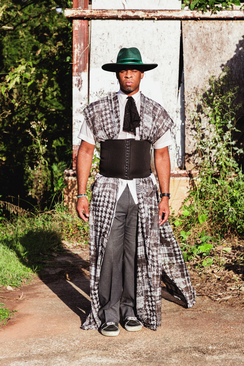 A man dressed in a fashionable, eclectic outfit standing outdoors. He wears a wide-brimmed green hat, a black corset over a white shirt, patterned fabric draped over his shoulders, gray pinstripe pants, and black sneakers. He is standing on a dirt pa