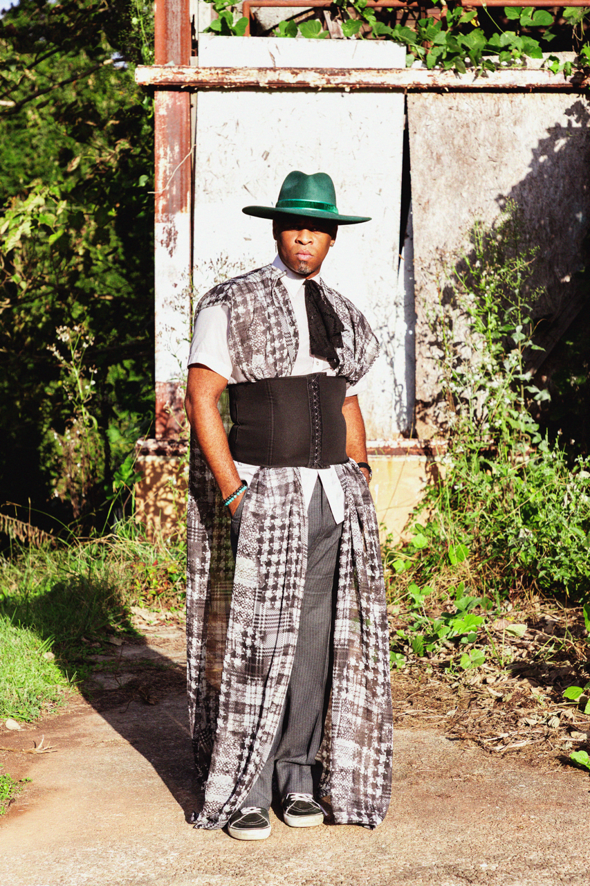 A person standing outdoors in front of a weathered wall, wearing a large green hat, a patterned long coat, wide pants, and a black corset over a white shirt, with greenery around.