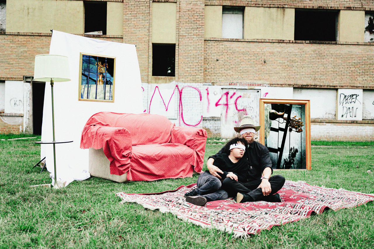 A man and a child sit on a patterned rug outdoors in front of a dilapidated building. The child is blindfolded and resting on the man's shoulder. The scene includes a red-covered sofa, a standing lamp, and framed artworks leaning against the building