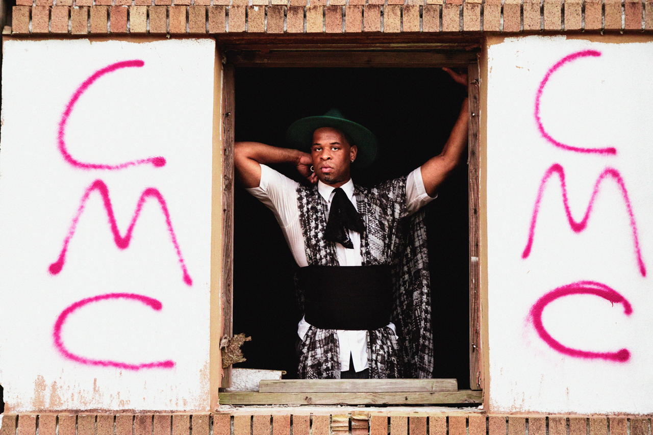 A man dressed in vintage Western-style clothing, wearing a large hat, standing in a window with graffiti that spells 'C M C' on white walls on either side.