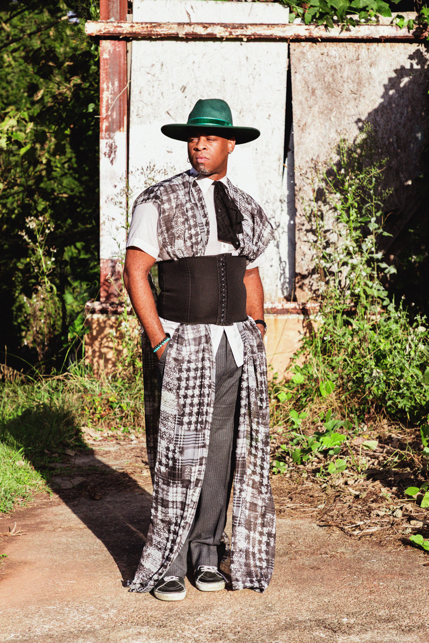 A person standing outside in front of a weathered wall, wearing a wide-brimmed green hat, patterned outfit with a corset, and casual shoes, with greenery around.