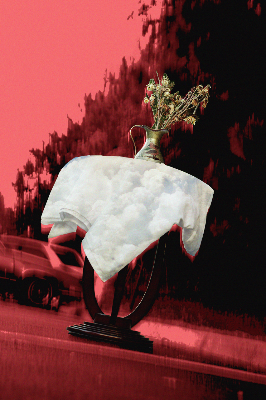 A decorative sculpture featuring a white T-shirt with clouds printed on it, draped over a black circular stand, with a brass vase holding dried flowers placed on top, set against a pink and black abstract background with a blurred car passing by.