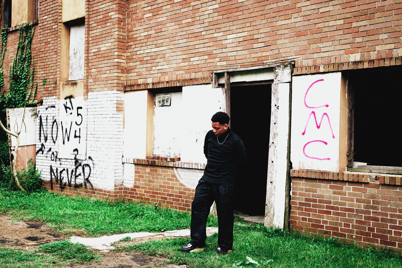 Young man wearing black hoodie and pants standing outside a brick building with graffiti on the walls.