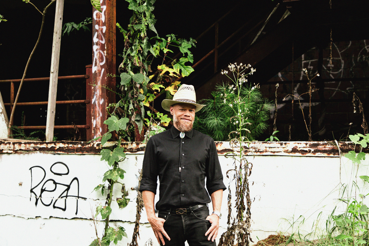 A man with a beard wearing a white cowboy hat, black button-up shirt, and black jeans standing outdoors in front of a white wall with graffiti and overgrown plants.