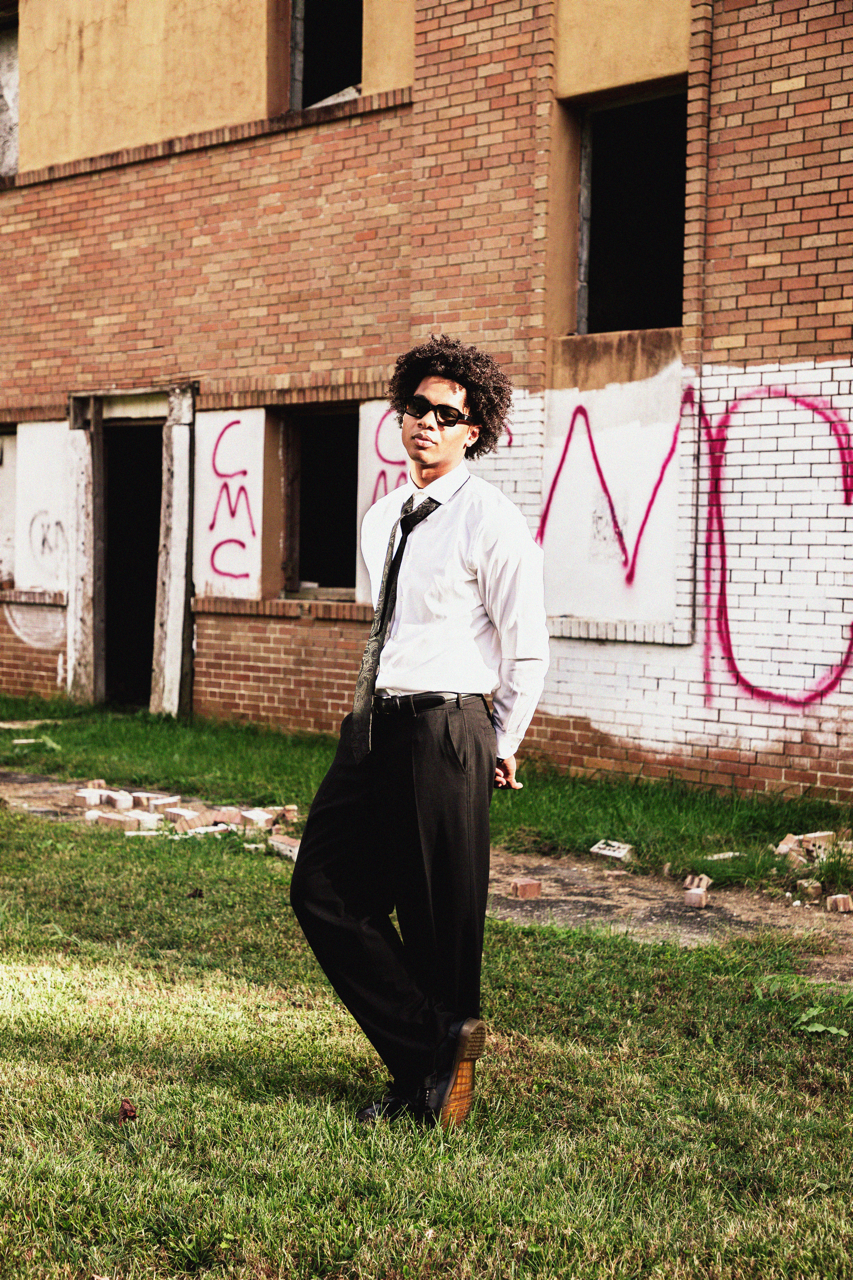 A man with curly hair wearing sunglasses, a white shirt, and a black tie, standing on grass in front of an abandoned brick building with graffiti.