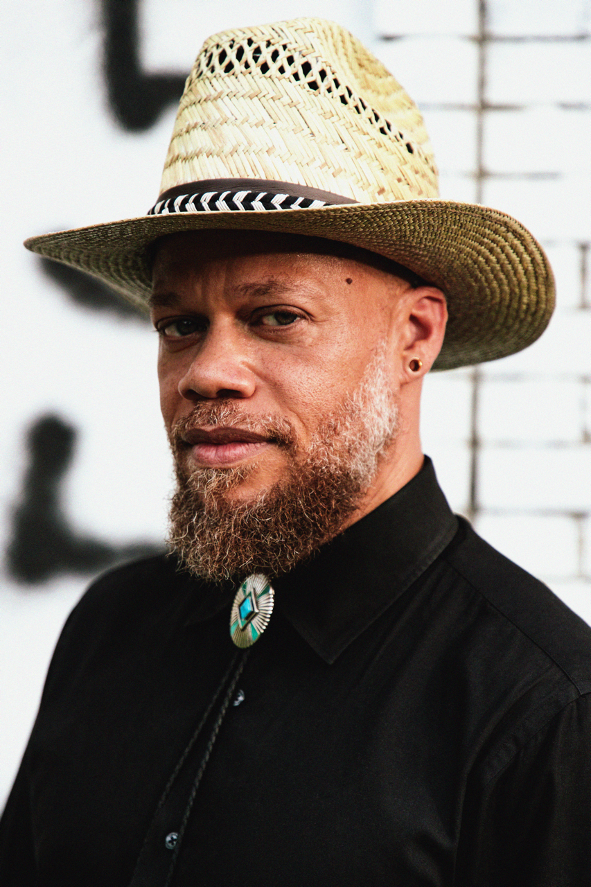 A man with a beard wearing a straw hat and a black shirt with a turquoise bolo tie, standing in front of a white brick wall with black abstract art.