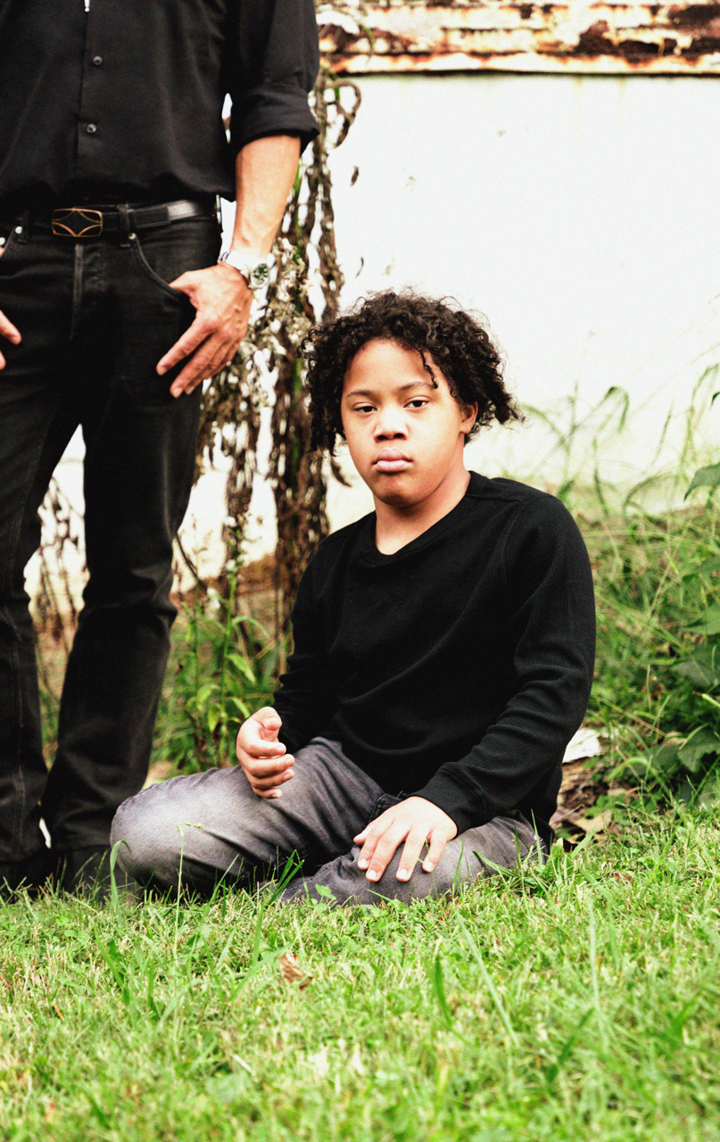 A young boy with curly hair sitting cross-legged on grass next to a person standing, partially visible. The boy is wearing a black long-sleeve shirt and gray pants, with a serious expression. The setting appears to be outdoors near a body of water an