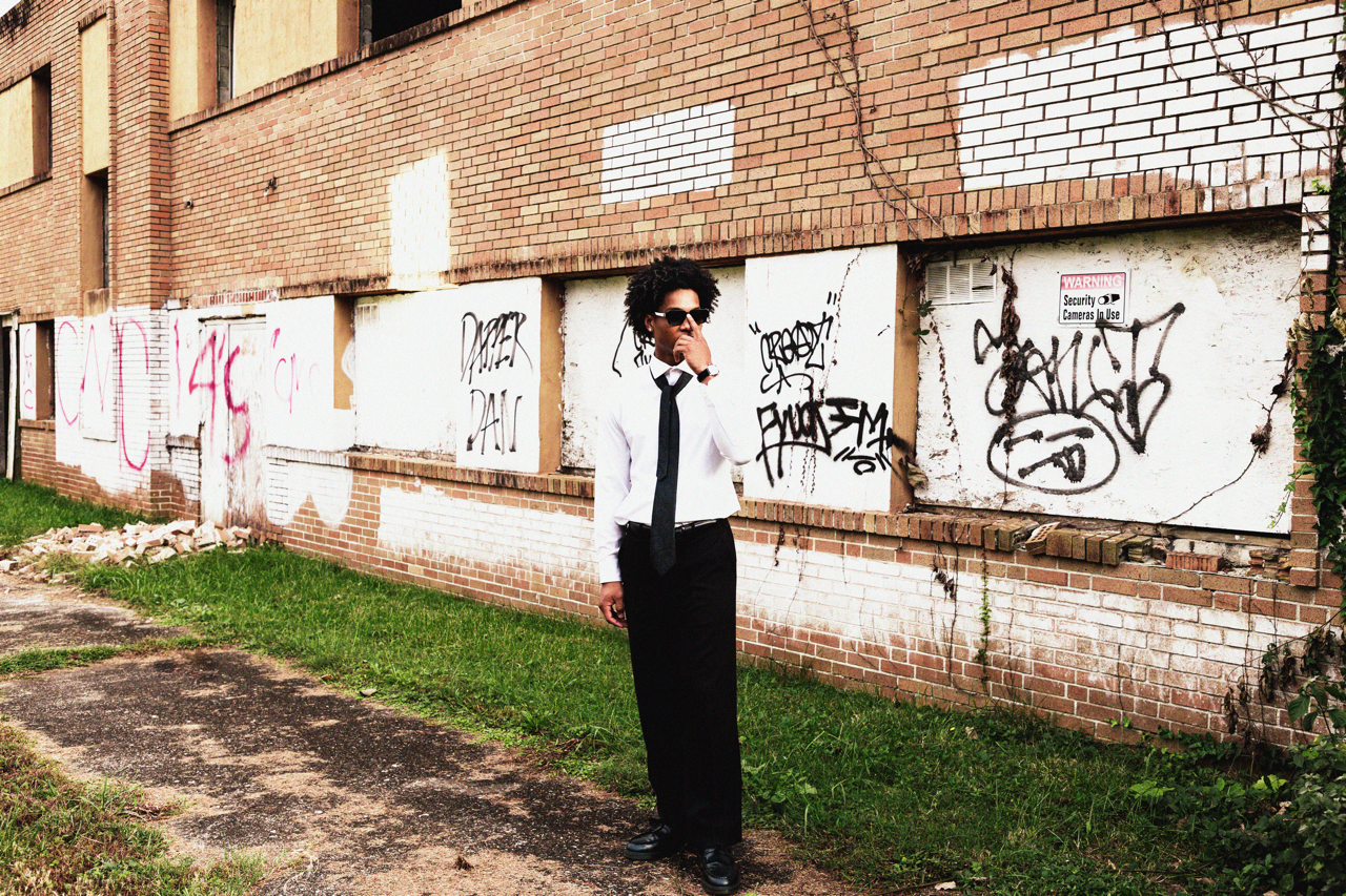 A woman in a white shirt, black tie, and long black skirt standing in front of a brick wall with graffiti. She is wearing sunglasses and holding a cigarette near her mouth.