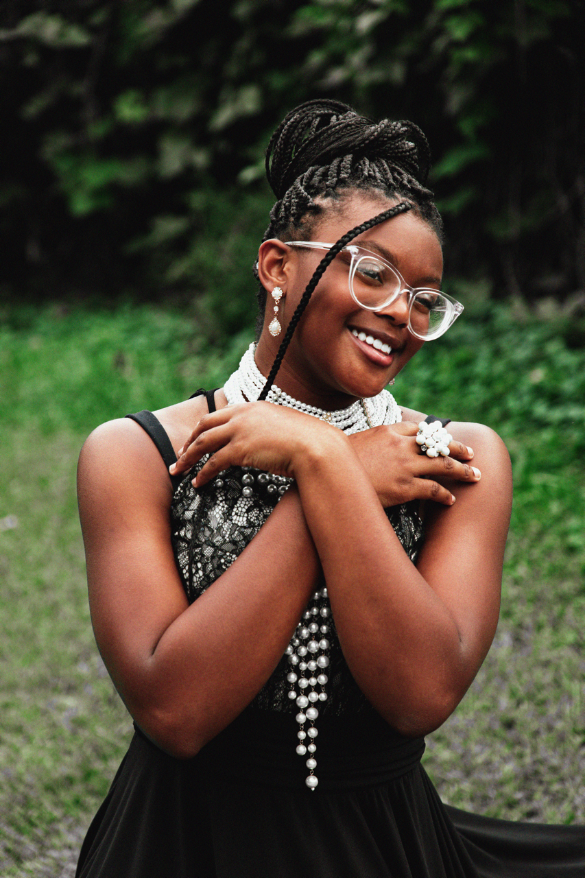 A smiling woman with dark skin and braided hair styled in an updo, wearing large clear glasses, pearl earrings, layered pearl necklaces, a ring, and a black dress with lace details, standing outdoors with greenery in the background.