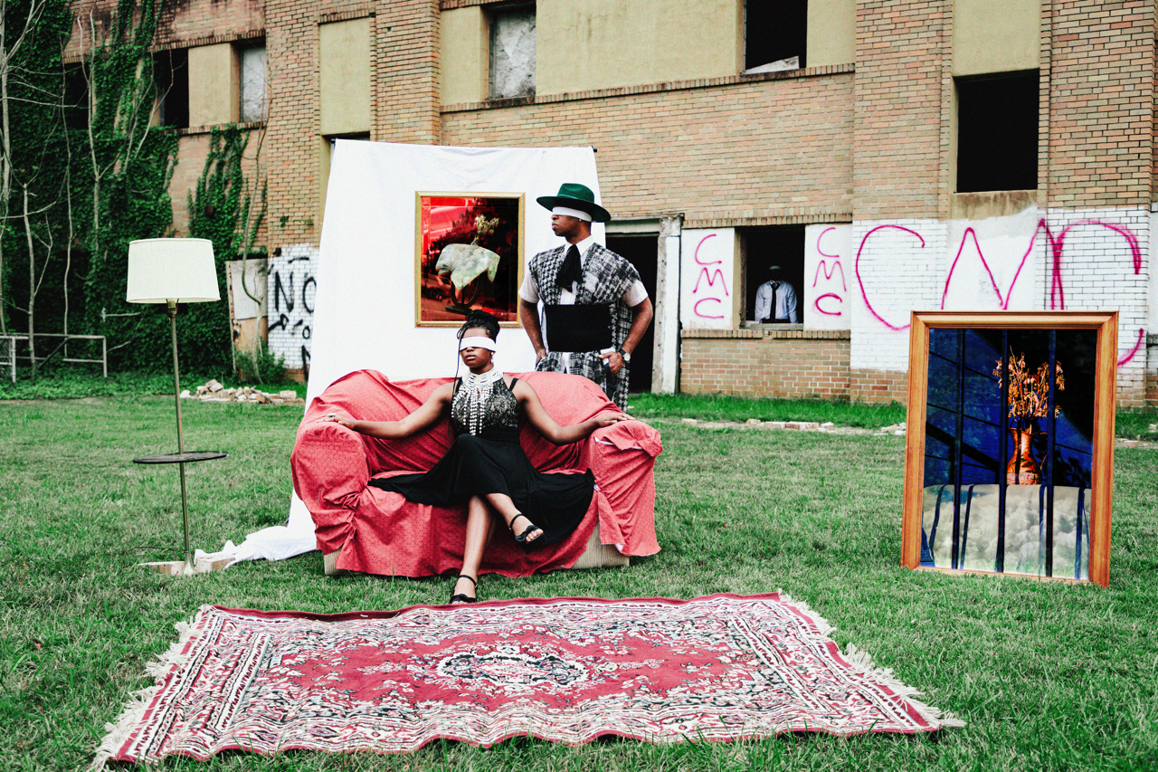 A woman with a blindfold sitting on a pink couch outdoors with a man standing behind her. The scene is set on a grassy area with a rug, a floor lamp, and framed pictures, and an unfinished brick building with graffiti in the background.