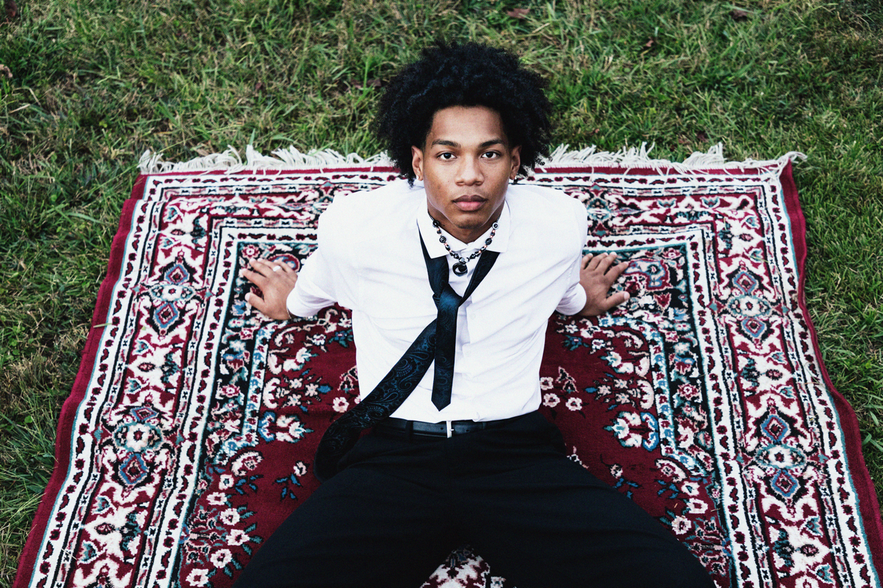 A young man with curly black hair sitting on a patterned rug outdoors on grass, wearing a white shirt, black pants, and a loosened black tie, looking up at the camera.