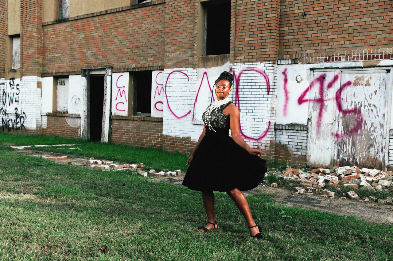 A woman in a black dress and high heels standing on grass in front of a partially demolished brick building with graffiti on the walls.