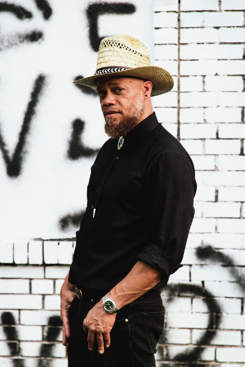 A man wearing a straw hat, black shirt, and black pants standing against a graffiti wall with white bricks.
