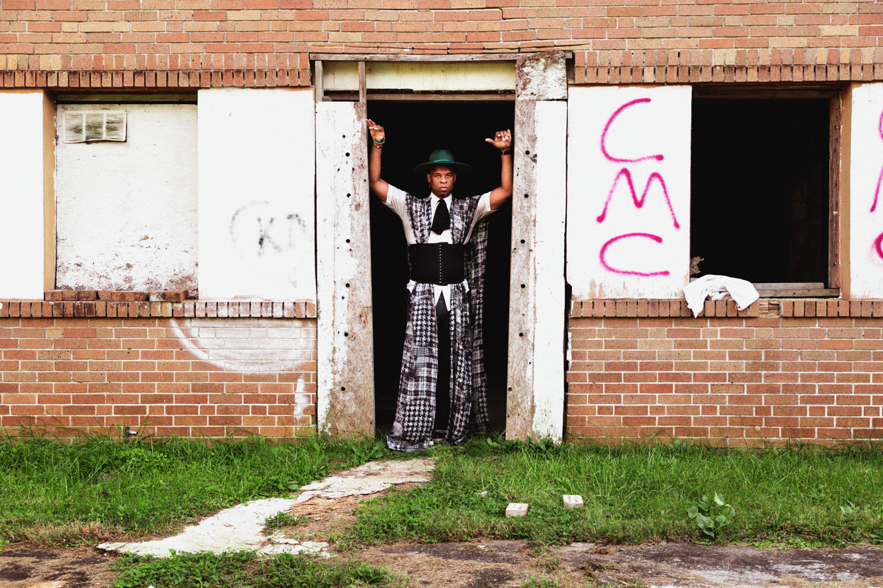 A person standing in a doorway of an abandoned brick building with broken windows, graffiti on the walls, and a white cloth draped over the sill, wearing a wide-brimmed hat and patterned clothing.