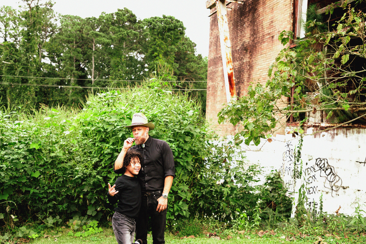 A man and a boy standing outdoors near greenery and graffiti on a wall, with the man smoking a cigarette and the boy making a peace sign.