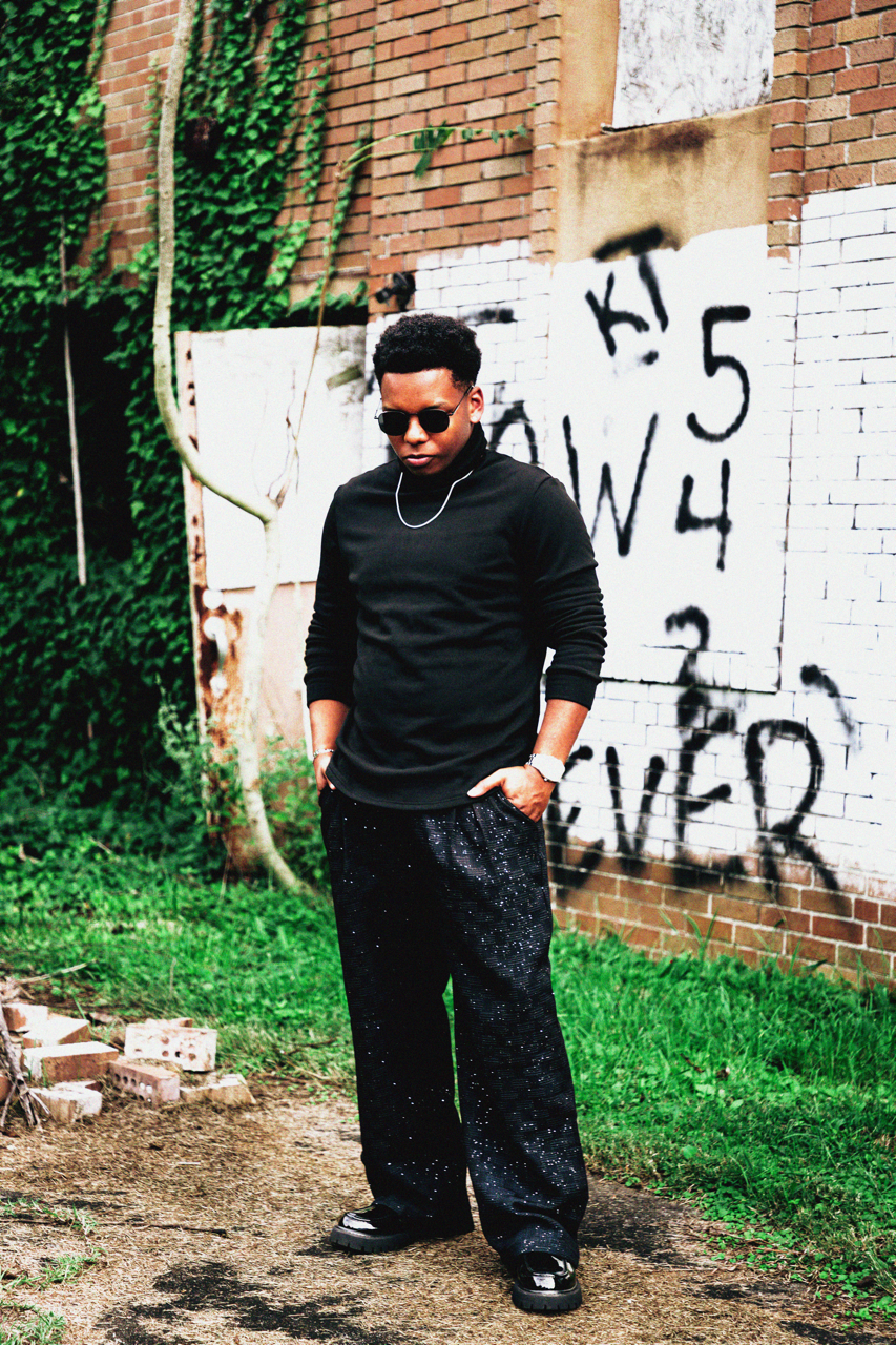 A young man with dark skin, wearing black sunglasses, a black long-sleeve shirt, and black pants with white speckles, embracing a street fashion style, stands with his hands in his pockets in front of a brick wall with graffiti and greenery.