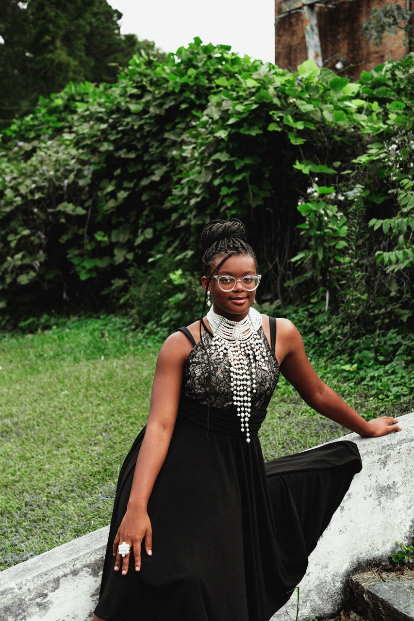 A woman in a black dress with intricate lace and an elaborate pearl necklace, standing outdoors on a concrete ledge with lush greenery behind her.
