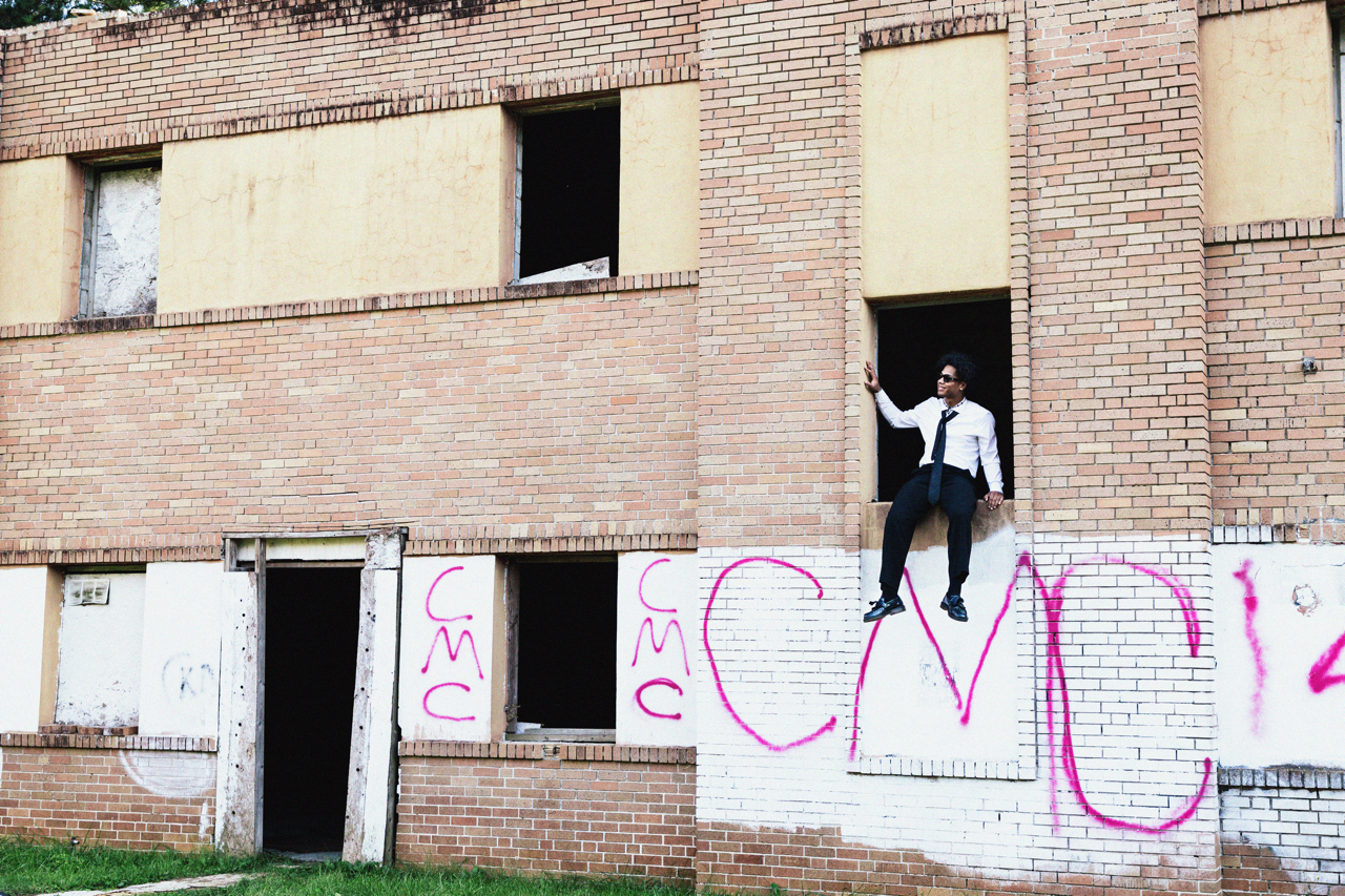 A man in formal attire, wearing sunglasses, sitting on a ledge in front of an unfinished brick building with graffiti on the lower wall.