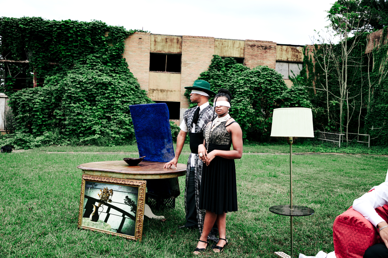 Two individuals wearing blindfolds stand beside a table with a framed painting leaning against it, in an outdoor setting with green grass and overgrown bushes and a partially dilapidated brick building in the background. One person is touching the ta