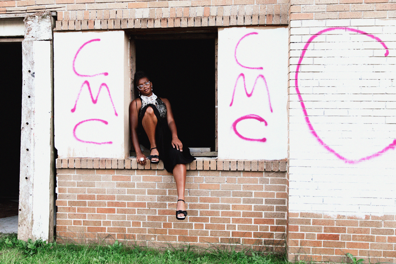 A woman in a black dress and glasses sitting in an empty window frame of a brick building with pink spray-painted graffiti on the walls around her.