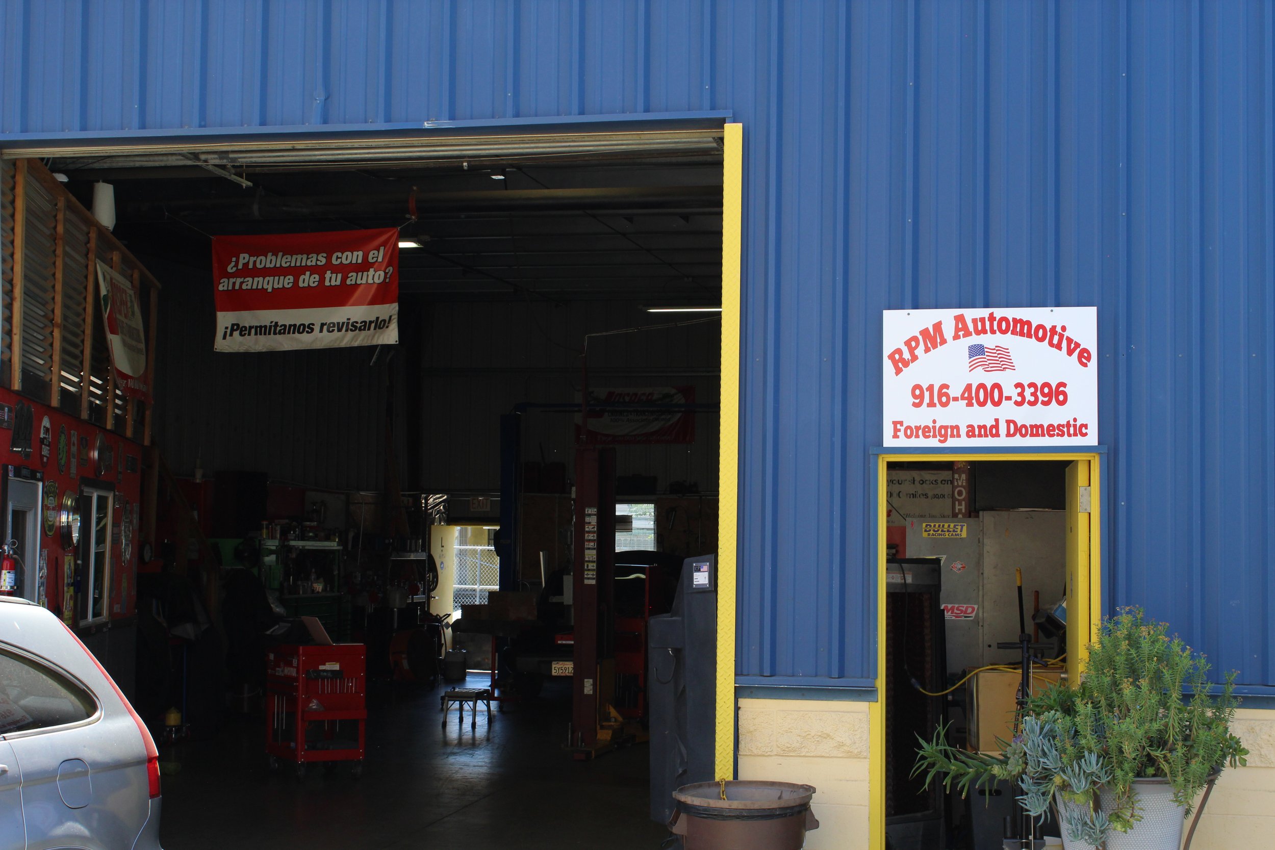 Exterior of an auto repair shop with a blue building, yellow trim, and a sign that reads 'RPM Automotive, 916-400-3396, Foreign and Domestic.' An open garage door reveals a dark interior with equipment and car repair tools. There is a small plant in 