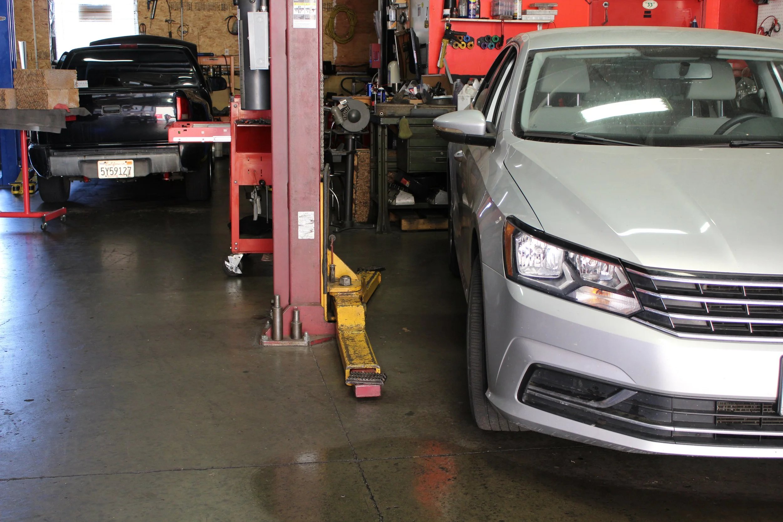 Inside an auto repair garage with a gray sedan on the right and a black vehicle on the lift in the back, surrounded by tools and equipment.