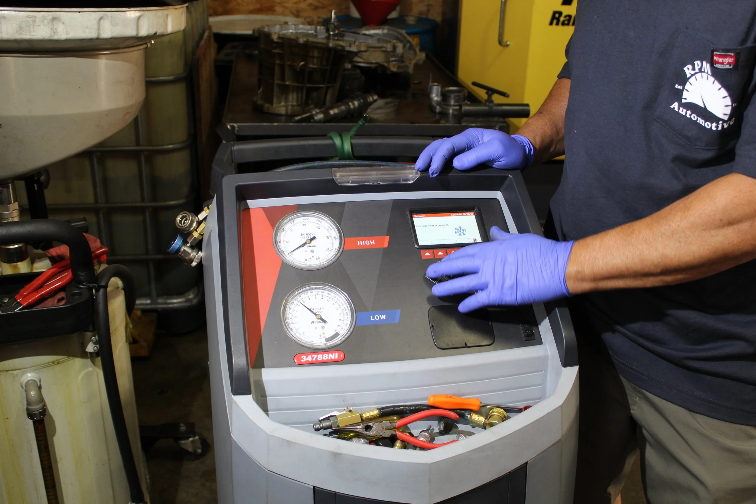 A person wearing blue gloves operating an automotive air conditioning service machine in a workshop.